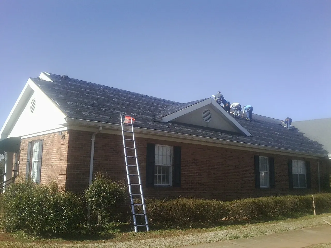Crew tearing shingles off a brick ranch roof with a ladder set — Emergency Roof Repair work by our crew in Sanders