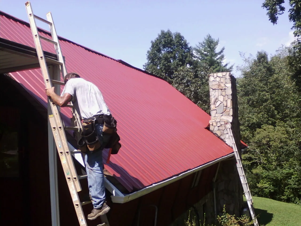 Roofer on a ladder beside a red metal roof and stone chimney — Soffit & Fascia Repair by Roofing Brothers in Sanders